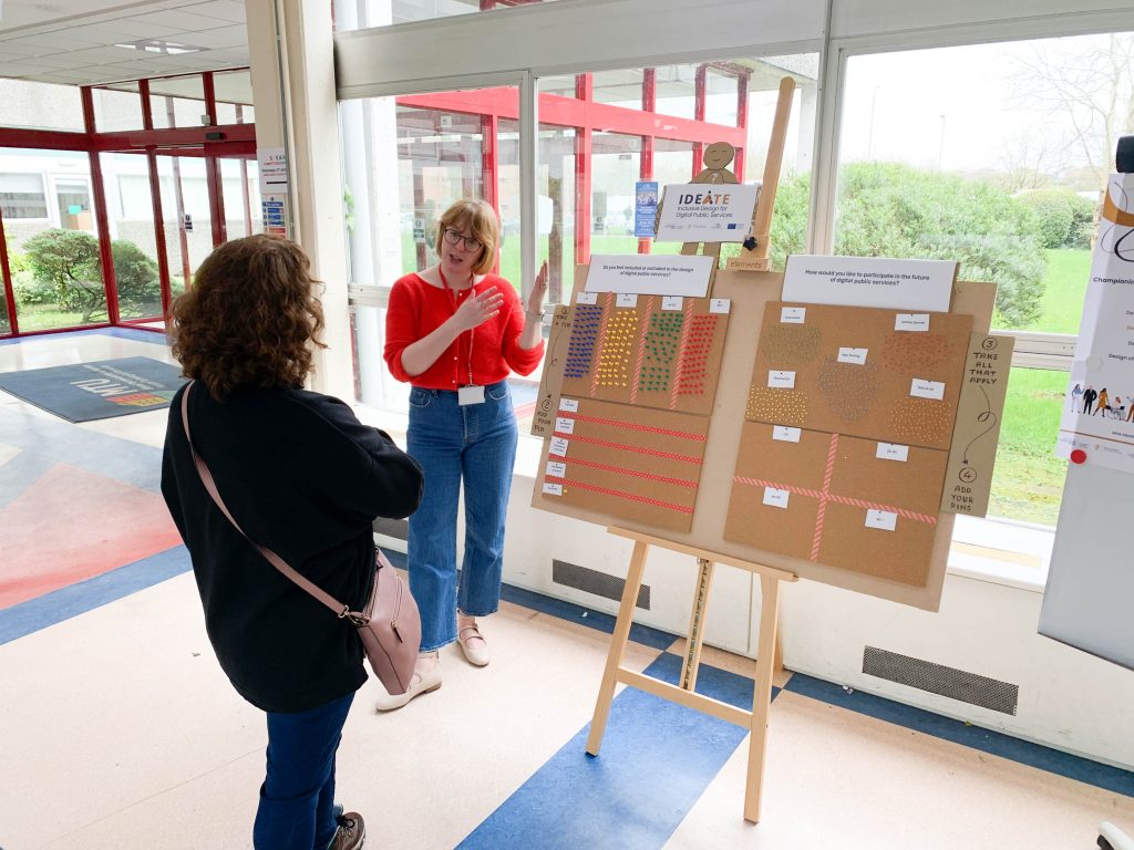 A women wearing a red jumper and jeans is discussing and gesturing towards a Data Board, while another woman wearing black is listening with her arms crossed